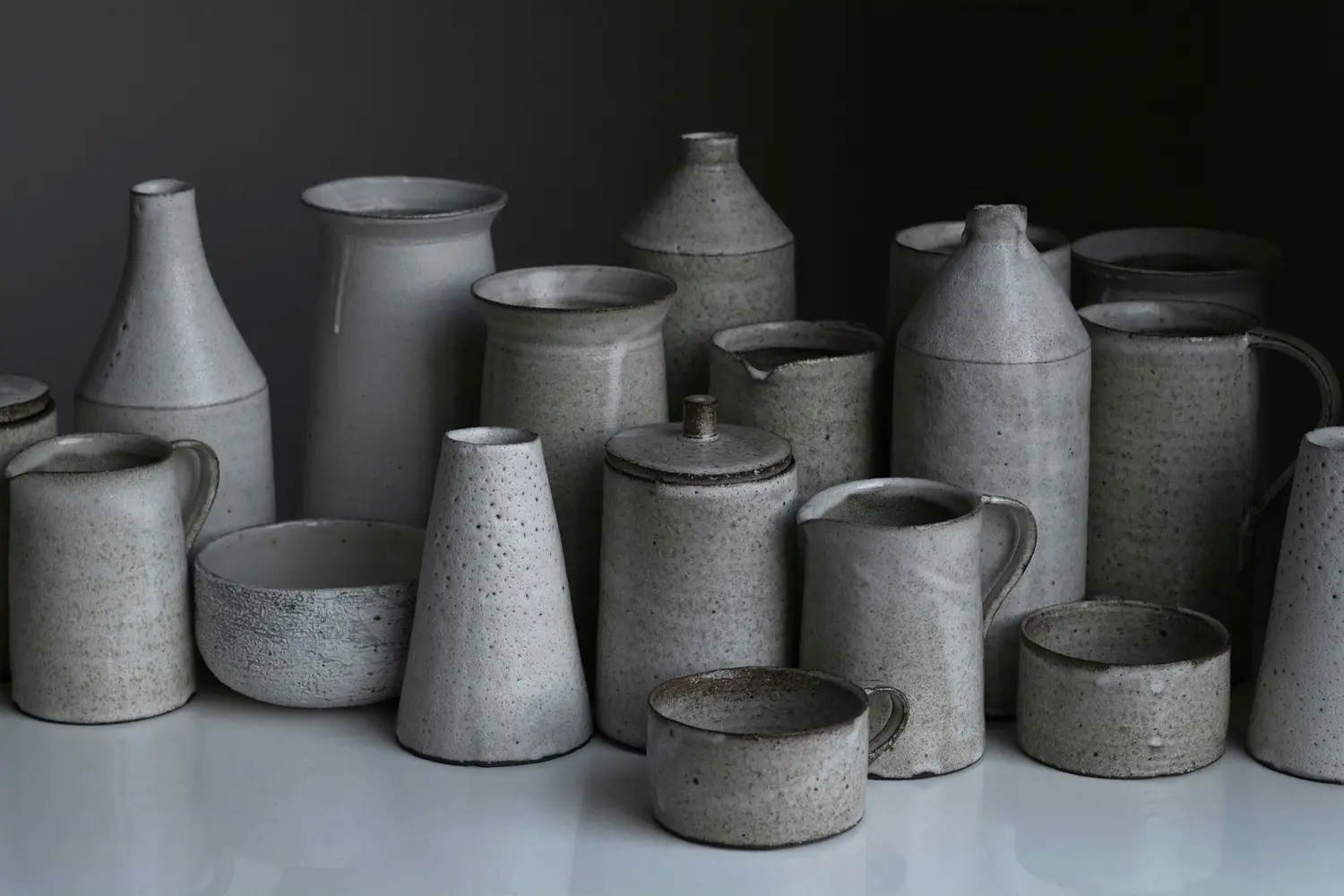 Studio table with speckled white stoneware jugs, bottles and bowls against a dark grey wall